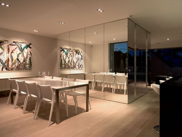 A sophisticated dining room with a long rectangular dining table and white chairs, set against a mirrored feature wall. Warm recessed lighting, pale timber flooring, white cabinetry or sideboard surfaces, and abstract artwork create a refined contemporary interior. Full-height glazing or reflective panels open toward a darker adjoining area outside the main dining space.
