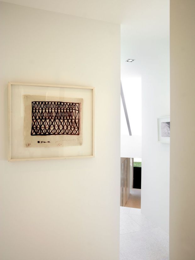 A bright minimalist interior view from a narrow passage or entry area, with a framed geometric artwork mounted on a warm off-white wall. An opening leads toward a sunlit room with pale flooring, white walls, a recessed ceiling light, and glimpses of timber joinery and outdoor greenery.