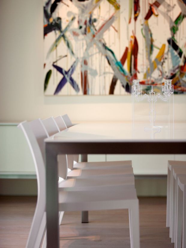 A bright, refined dining room centered on a long dark wood dining table with white modern chairs. A transparent decorative candelabra sits on the tabletop, while a large colourful abstract artwork forms the backdrop. Pale walls, simple trim, and warm timber flooring create a clean contemporary setting.