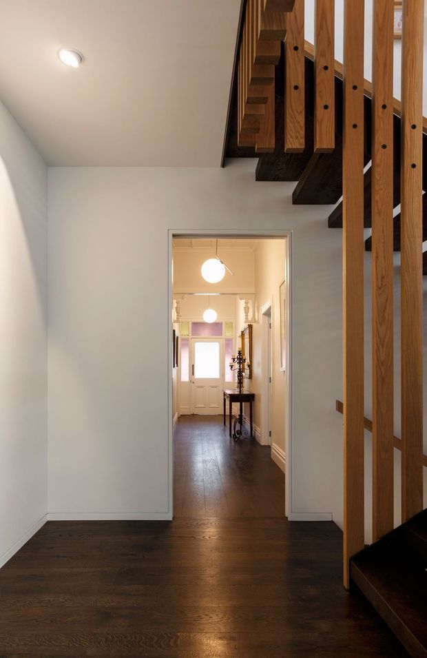 Interior view of a residential hallway and stairway. The foreground has dark timber flooring and a contemporary open-riser timber staircase with vertical timber balustrade slats. A white-painted passage leads to a traditional entry hall with a glazed front door, console table, wall mouldings, and pendant lights.