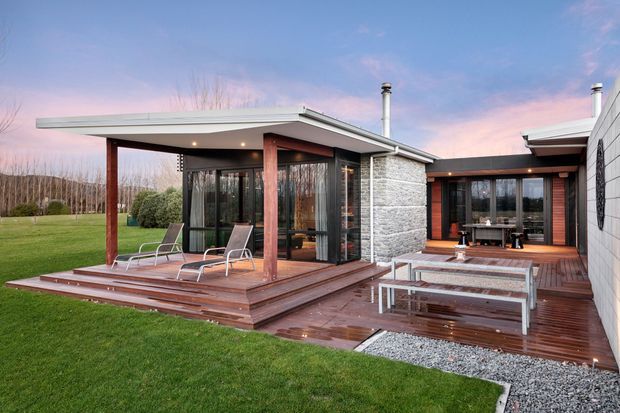 Single-storey contemporary home arranged around a timber-decked outdoor entertaining area. The low-pitched roof, extensive glazing, dark timber posts and cladding, and prominent textured masonry feature wall create a modern indoor-outdoor setting. The foreground includes a stepped timber deck with outdoor chairs, a low table, and a manicured lawn; the background shows a wooded landscape at dusk.