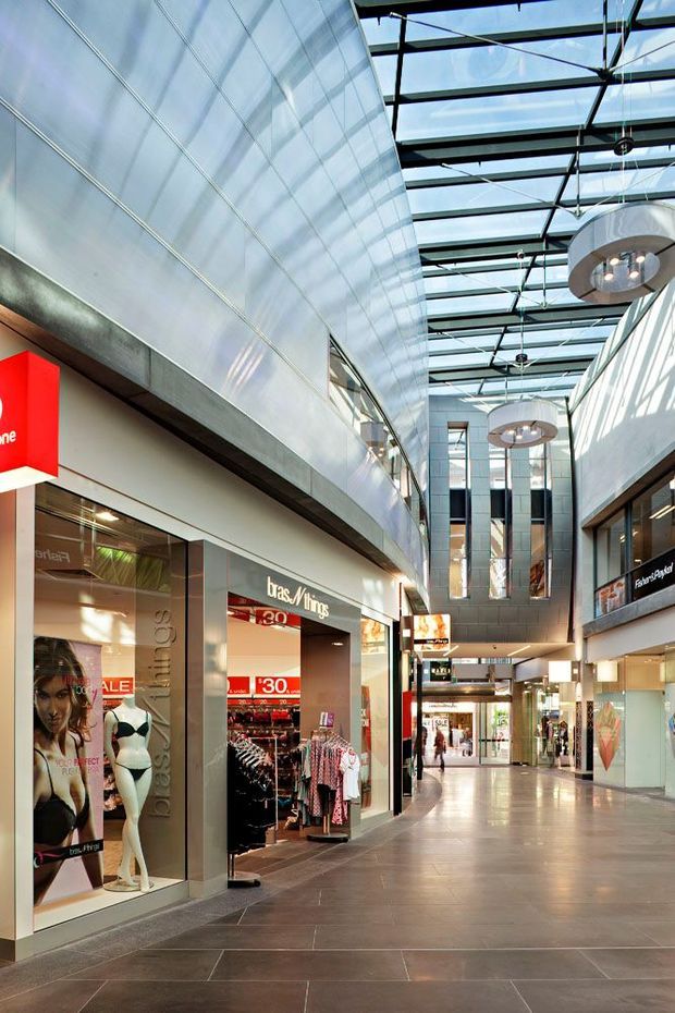 Interior view of a contemporary multi-level shopping mall corridor, with retail storefronts on both sides, a tall glazed atrium roof, exposed dark structural framing, large pendant lights, and polished tiled flooring. Bright daylight enters through the roof and upper-level glazing; visible signage and clothing displays indicate a commercial retail environment.