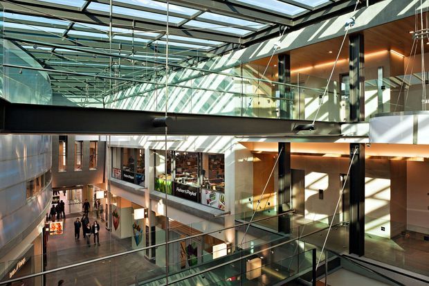 Interior view of a modern multi-level commercial atrium or shopping complex, featuring extensive skylights, glass balustrades, exposed dark structural framing, suspended pendant lights, and retail storefronts around an open circulation space. Strong daylight creates geometric shadows across the walls and floors.