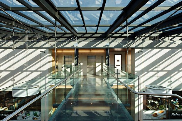 A bright contemporary commercial interior viewed from an elevated glass walkway or mezzanine. The space features a transparent glazed roof, exposed dark steel beams and services, extensive glass balustrades, polished reflective flooring, and strong geometric sunlight and shadow patterns. Retail or display areas are visible on the lower level at the edges.