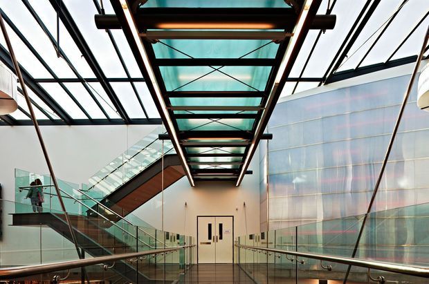 A spacious contemporary interior circulation space with a central glazed overhead walkway or skylight structure, exposed dark steel framing, glass balustrades, and a prominent translucent wall. A stair and escalator-like circulation route rise on the left, while a polished reflective floor and metal handrails lead toward double doors. The space has strong linear geometry, abundant daylight, and a commercial or institutional character.