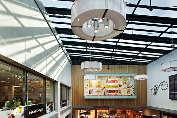 A bright, multi-level commercial interior with a dramatic glazed roof structure, exposed dark steel framing, and oversized circular pendant lights. The space features a tall timber-lined feature wall, a colourful illuminated display or retail counter, extensive glazing, and visible lower-level commercial activity. The overall setting appears contemporary and highly designed, with strong daylight and an atrium-like volume.