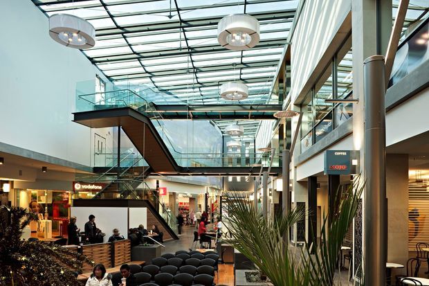 Interior view of a multi-level contemporary commercial shopping and dining atrium, with a glazed roof, extensive internal glazing, suspended circular pendant lights, escalators, retail storefronts, cafes, seating, and indoor planting. The space has a bright, open public-complex character with visible upper-level walkways and exposed structural framing.