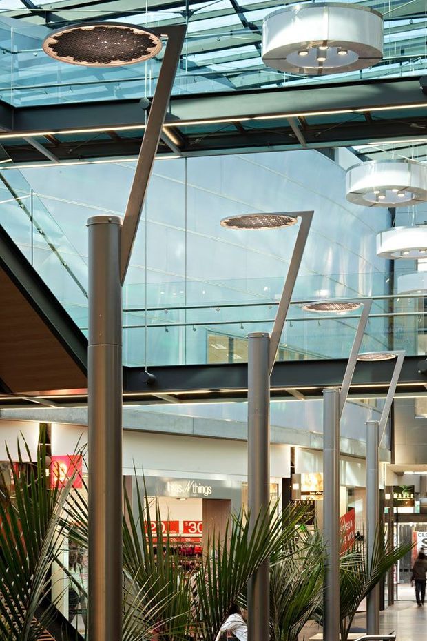 Interior view of a large, bright commercial shopping or retail atrium with extensive glass roof and walls, exposed dark structural beams, suspended circular ceiling lights, and a row of tall sculptural light poles. Indoor tropical planting and retail storefronts are visible at ground level, creating a contemporary public mall environment.