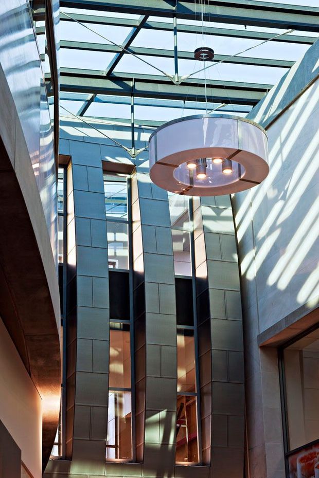 A tall contemporary interior atrium or lobby with a glazed roof, exposed dark structural beams, extensive vertical glazing, and reflective metal wall panels. A large suspended circular pendant light hangs centrally, while strong sunlight creates geometric shadows across the walls. The space has a dramatic multi-level vertical volume.