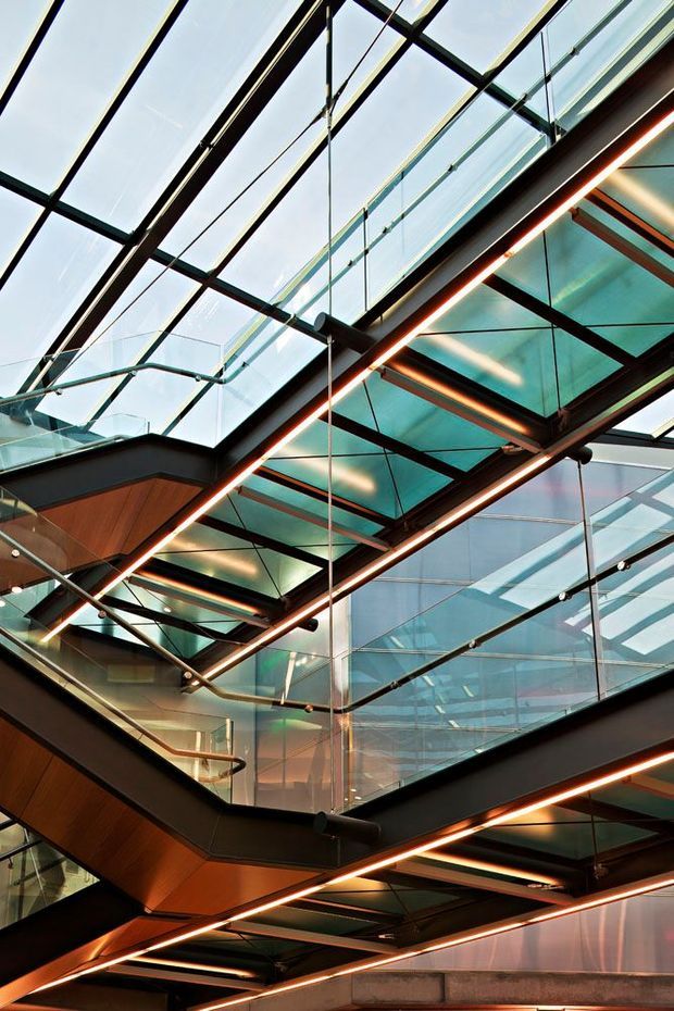 Upward-looking view inside a large contemporary commercial atrium, showing layered steel-framed glazed walkways or stair structures beneath a transparent roof. Exposed dark structural beams, glass balustrades, warm timber soffit panels, and linear integrated lighting create a dramatic high-tech interior.