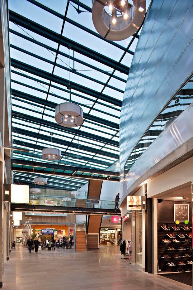 Interior view of a contemporary multi-level retail shopping centre or commercial arcade. The spacious corridor has polished large-format flooring, retail storefronts, a mezzanine walkway, a dramatic glazed roof structure with exposed dark framing, suspended circular pendant lights, and metallic wall cladding. Shoppers and shop displays are visible in the distance.