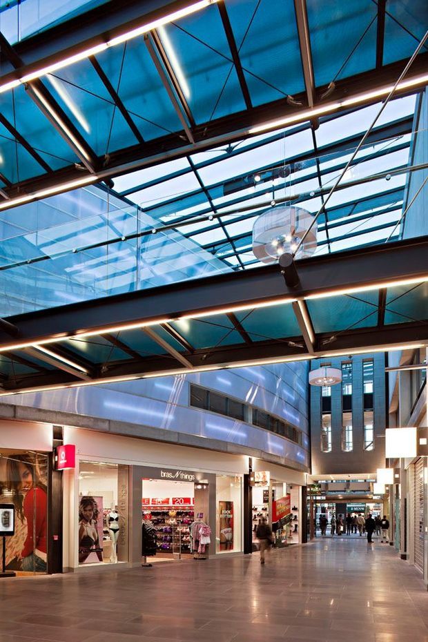 Interior view of a contemporary multi-level shopping mall or commercial arcade, with retail storefronts lining a broad polished floor corridor. The space features a dramatic glazed atrium roof, exposed dark steel beams and braces, suspended linear lighting, large ceiling fans, reflective metal wall cladding, and a tall vertical circulation or atrium zone in the background. Bright retail signage and shoppers are visible, but the dominant architectural character is the expansive commercial interior and overhead glazing.