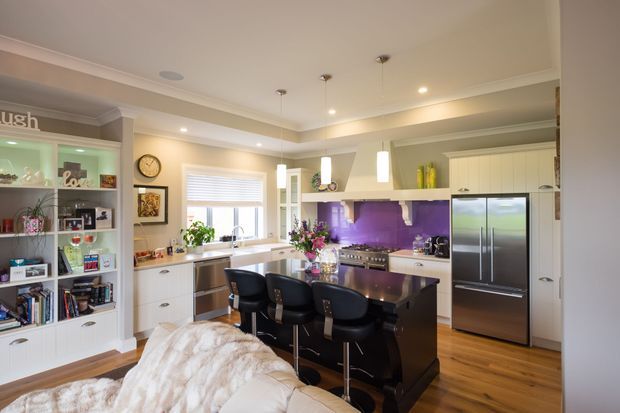 Bright, spacious open-plan kitchen viewed from an adjoining living area. The room features white shaker-style cabinetry, a dark central island with seating for four, stainless-steel appliances, a vivid purple splashback, timber flooring, pendant lights, and integrated open shelving. A sofa is visible in the foreground, confirming the kitchen connects directly to the living space.