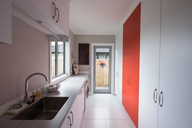 A narrow, modern galley kitchen with a long pale benchtop, stainless-steel double sink and curved tap beneath a window. White and pale pink cabinetry lines the room, with a bold red full-height cabinet wall providing a strong feature accent. Large light floor tiles, a rear glazed door, and compact integrated storage complete the space.
