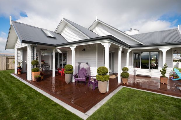 Exterior view of a large single-storey white weatherboard home with multiple pitched gables, grey metal roofing, and a broad covered veranda opening onto a polished timber deck. The deck is furnished with outdoor seating and potted topiary, with manicured lawn surrounding the house.
