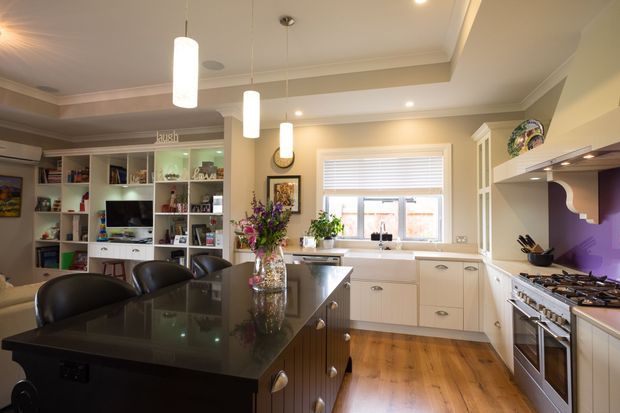 A bright, traditionally styled kitchen viewed from the dining or adjoining living area. The room features a large dark stone island with seating, white shaker-style cabinetry, timber flooring, a tiled or painted backsplash area, extensive built-in shelving, pendant lights, a gas range, and a window above the sink. Decorative shelving and floral arrangements add a homely, refined character.