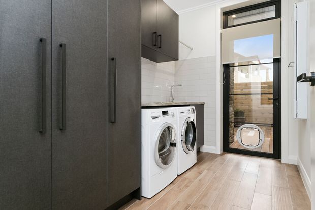 A compact, modern laundry room with dark full-height storage cabinetry, white front-loading washer and dryer, white subway-tile walls, and warm timber-look flooring. A glazed black-framed door opens to an outdoor area, while upper cabinetry provides additional storage.