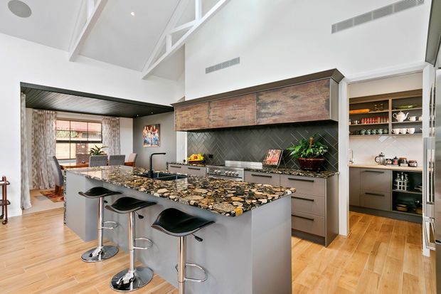 A bright, open-plan contemporary kitchen with a large waterfall-edge island, four black bar stools, integrated cooking appliances, dark herringbone tile splashback, grey cabinetry, and distinctive rustic timber overhead cabinetry. The kitchen opens toward a dining or living area and features a high pitched ceiling with exposed white structural framing.
