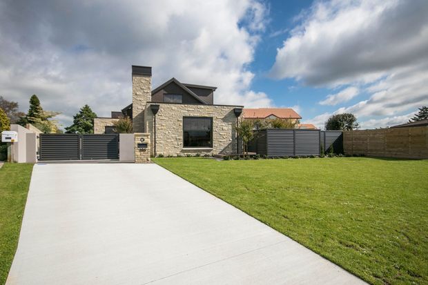 Street-facing view of a contemporary single-storey residential facade with a prominent light stone-clad wall, dark charcoal metal fencing and garage gate, pitched roof forms, and a broad concrete driveway leading to a manicured front lawn. The house features a chimney-like stone element, dark-framed glazing, and a partially visible timber-clad boundary fence.