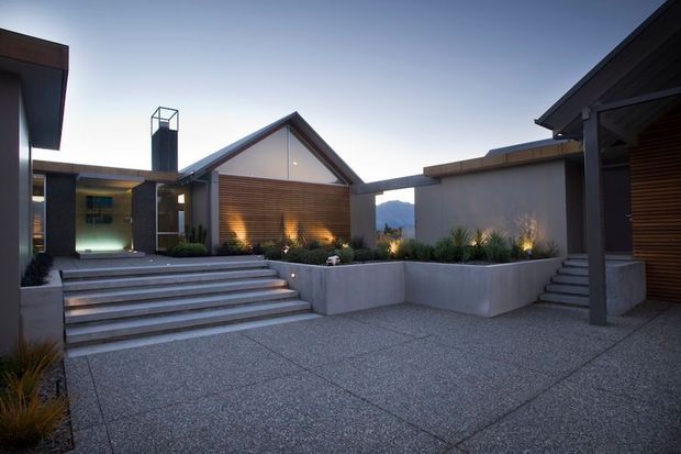 Twilight exterior view of a contemporary residential courtyard and entrance. The composition features multiple angular, gabled and flat-roofed volumes, broad concrete steps, a large textured paved forecourt, rendered concrete planter walls, dark metal framing, and warm horizontal timber screening illuminated from within. Sparse architectural landscaping and distant hills are visible beyond the courtyard.