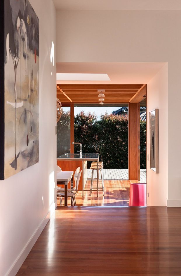 A warm, sunlit interior view looking down a hallway into an open-plan kitchen and dining area. Dark timber flooring leads to a compact kitchen with timber cabinetry and a stainless-steel benchtop, while a timber-framed glazed opening connects to a deck and leafy outdoor area. The kitchen ceiling features a distinctive timber slatted treatment with pendant lighting, and white walls display large abstract artwork. A bright red bin provides a strong accent.