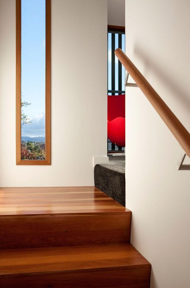 Interior photograph of a contemporary stairway with warm timber treads and landing, a simple timber handrail, white plastered walls, and a narrow timber-framed vertical window overlooking a landscape. The upper stair area includes dark carpet and a bold red feature visible beyond the opening.