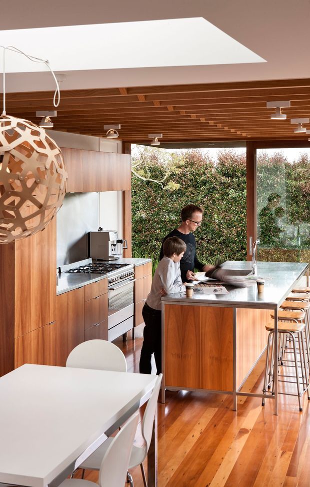 Bright, open-plan kitchen and dining area with extensive warm timber cabinetry, a large white island with integrated sink and seating, stainless-steel appliances, white dining furniture, and floor-to-ceiling glazing overlooking greenery. The ceiling features exposed timber slats and recessed linear lighting, while a sculptural pendant hangs above the dining area. Two people are gathered at the island, reinforcing the family-oriented kitchen setting.