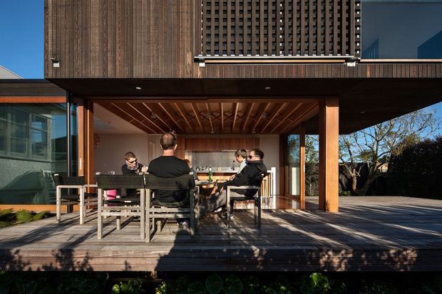 Contemporary timber-clad residential extension with a covered outdoor dining deck opening directly into a bright kitchen and living area. The image features strong indoor-outdoor connection, exposed timber ceiling battens, large glazed openings, timber posts, and built-in cabinetry, with people seated around an outdoor dining table.