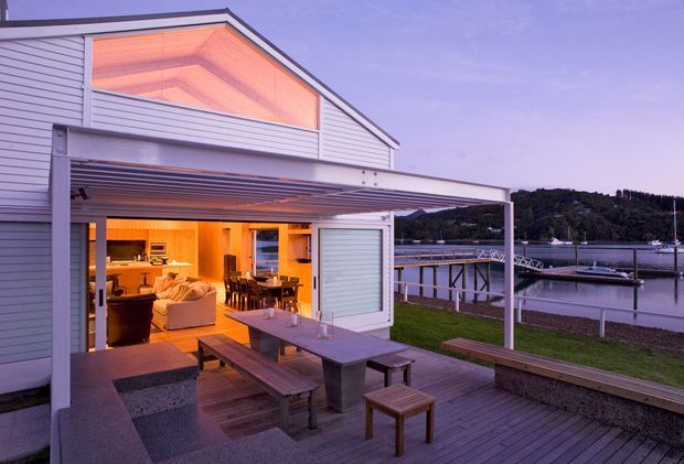 A contemporary waterfront home photographed at dusk, with a light horizontal-clad gabled facade and a large covered outdoor entertaining deck. Open glazed doors connect the deck to a warmly lit open-plan interior containing a kitchen, dining area and living room. Timber decking, built-in concrete seating, picnic-style dining furniture and a broad pergola frame the harbour and private jetty beyond.