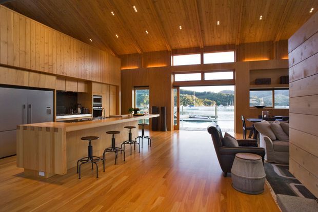 Open-plan timber-lined kitchen and living area with a large central island, integrated appliances, bar stools, lounge seating, and expansive glazed doors and clerestory windows opening toward a calm waterfront view. Warm recessed lighting and extensive natural timber create a refined, inviting interior.