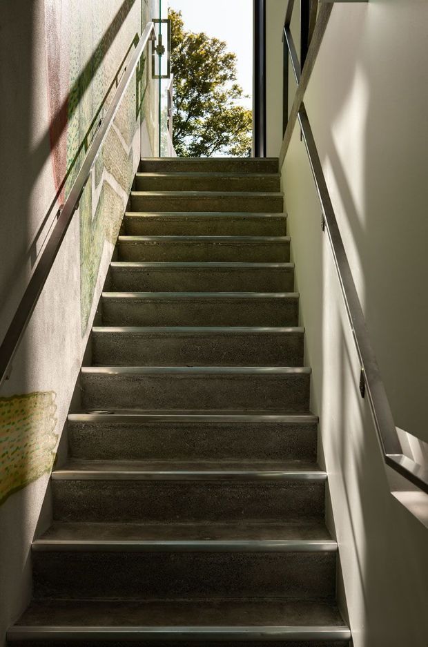Interior view looking up a narrow stairway with dark textured concrete or stone steps, slim metal handrails on both sides, pale walls, and strong sunlight entering through tall glazing at the landing. A leafy tree is visible outside, and an artwork or patterned wall covering appears along the left wall.