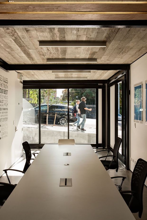A compact contemporary meeting room or office with a long white conference table, black office chairs, exposed dark structural beams, and a striking timber-plank ceiling with integrated linear lighting. Full-height glazed doors and windows provide views to the street, while white walls and black-framed glazing create a clean, industrial-leaning workspace.