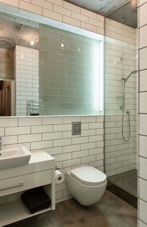 A compact contemporary bathroom with a wall-mounted white vanity and vessel basin, wall-hung toilet, large frameless mirror, and glass-enclosed walk-in shower. The walls are clad in white subway tiles with dark grout, while the shower features muted green-grey subway tiles. The exposed concrete ceiling and grey floor add an industrial character, with a vertical illuminated mirror edge providing a bright highlight.
