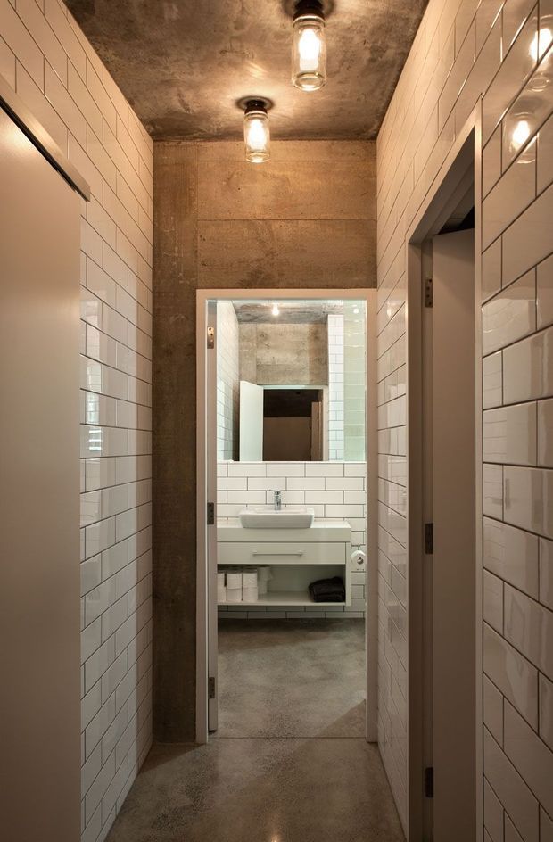 Narrow interior corridor leading into a compact bathroom. Glossy white subway tiles line both side walls and continue around the vanity area, contrasted with raw concrete walls and ceiling. The bathroom contains a white wall-mounted vanity with an integrated basin, open lower shelving, and a large rectangular mirror. Exposed industrial-style pendant lights provide warm illumination, and a concrete floor reinforces the utilitarian aesthetic.