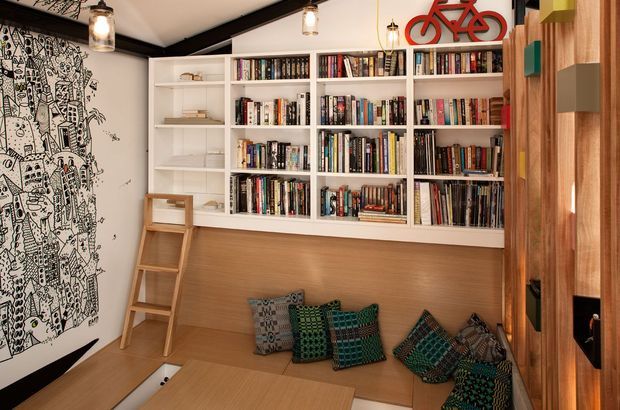 A compact, warmly lit reading nook or library corner with extensive white built-in bookshelves, a timber-lined lower bench, loose patterned cushions, and a timber ladder. A striking black-and-white illustrated wall mural occupies the left side, while vertical timber joinery with colourful open niches frames the right. Pendant lights and a red bicycle wall decoration add character to the eclectic space.
