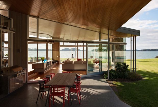 A mid-century-inspired open-plan dining and living area with a timber-lined angled ceiling, extensive floor-to-ceiling glazing, and strong indoor-outdoor connection. A timber dining table with red chairs occupies the foreground, while a living area and built-in cabinetry sit beyond. The glass walls open toward a broad lawn and waterfront landscape, with warm timber surfaces contrasting against dark flooring and black-framed glazing.
