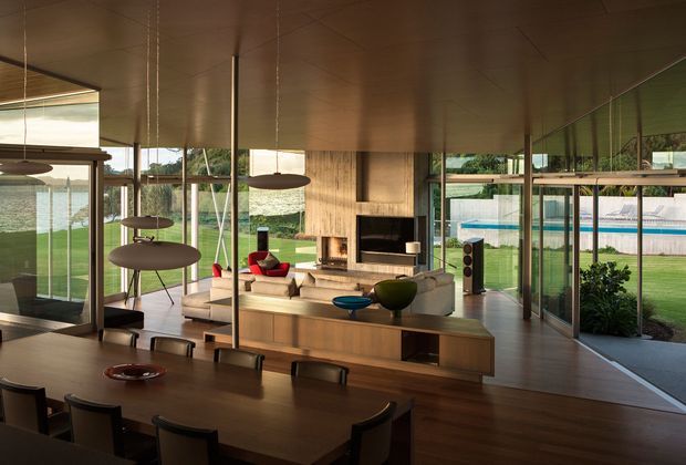 A spacious open-plan mid-century-inspired living and dining area with extensive floor-to-ceiling glazing overlooking a landscaped yard and swimming pool. The room features a long dark timber dining table, sculptural pendant lights, timber flooring and ceiling, a light upholstered sofa, a built-in timber-clad fireplace wall, and open-plan kitchen or joinery elements. The interior has strong indoor-outdoor connections and abundant natural light.