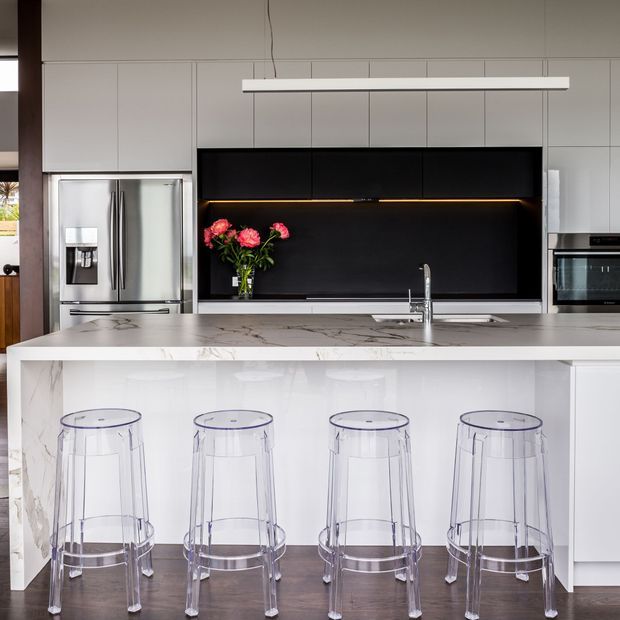 Contemporary kitchen featuring a large white marble island with waterfall edge, clear acrylic bar stools, white full-height cabinetry, stainless-steel appliances, and a dramatic black splashback with integrated warm lighting. A linear pendant light hangs above the island, while a vase of pink flowers adds colour.