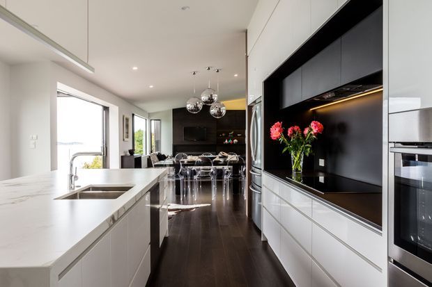 A bright contemporary kitchen with a long white island and integrated double sink, dark timber flooring, extensive white cabinetry, and a contrasting black feature wall with open display shelving and under-cabinet lighting. A stainless-steel refrigerator and built-in ovens line the right wall, while a dining area with reflective pendant lights sits beyond the kitchen. Large glazed openings bring in natural light and views to greenery.