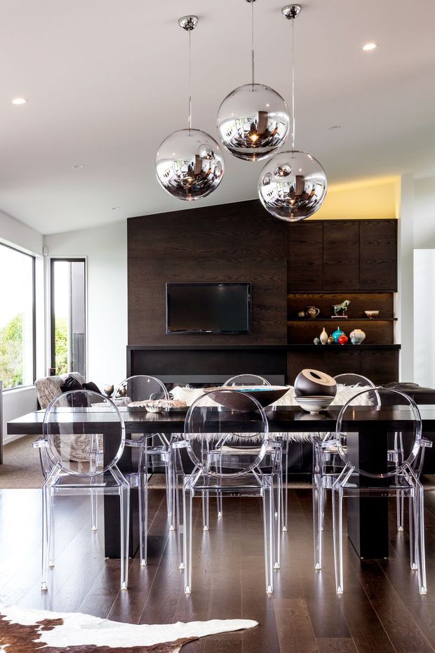 A sophisticated open-plan dining and living area with a dark timber feature wall and integrated cabinetry, black dining table, transparent acrylic dining chairs, and three reflective chrome globe pendant lights. Large windows bring in natural light, while decorative objects and a television occupy the built-in joinery. Dark timber flooring and a neutral palette create a polished contemporary interior.