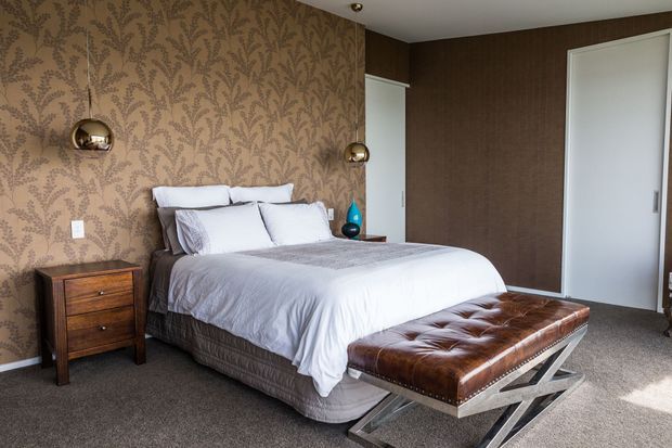 A well-appointed bedroom featuring a king-size bed with layered white and grey bedding, a tufted brown leather bench on a chrome X-frame base, dark wood bedside furniture, and warm patterned wallpaper. Wall-mounted spherical bedside lights and built-in sliding wardrobes complete the refined, vintage-inspired interior.