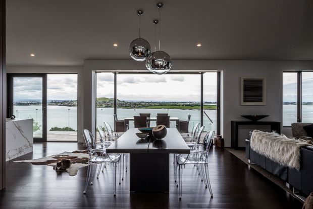 A refined open-plan dining and living area with a dark timber floor, pale walls, extensive floor-to-ceiling glazing and panoramic waterfront views. A long dark dining table is surrounded by transparent acrylic dining chairs, with sculptural chrome pendant lights overhead. A marble-look kitchen island is visible at left, while a dark sofa with a textured throw and a minimalist fireplace console occupy the right side.