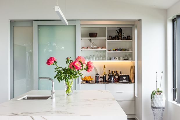Bright contemporary kitchen with a large white marble-look island, integrated sink and chrome tap, frosted-glass sliding pantry doors, and illuminated open shelving displaying glassware and kitchen appliances. A vase of flowers sits on the island, and daylight enters through a tall side window.