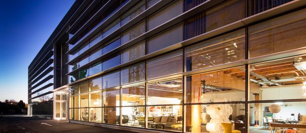 Twilight exterior view of a contemporary multi-storey commercial building with a long glazed ground floor and dark horizontal upper-level facade elements. Warmly illuminated interior workspaces, furniture, timber ceiling details, and exposed structural services are visible through the extensive curtain-wall glazing.