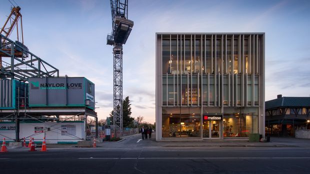 Street-level exterior view of a contemporary multi-storey commercial building with a tall vertical fins facade and extensive glazed frontage. Illuminated retail or hospitality interiors are visible at ground level, with a construction site, tower crane and temporary fencing to the left, and an adjoining low-rise commercial structure to the right at dusk.