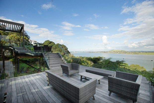 A spacious elevated timber deck overlooking a broad coastal bay or harbour. The outdoor living area contains woven lounge seating, a low coffee table and a built-in outdoor fireplace or fire pit, with stairs and glass balustrades connecting to a landscaped multi-level terrace. The setting is bright and relaxed, with expansive water views and contemporary architectural detailing.
