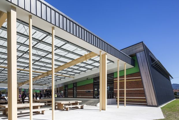Exterior architectural view of a contemporary public or commercial building with a large covered outdoor plaza. The structure combines red brick walls, vivid green panels, dark vertical metal cladding, extensive glazing, slender pale timber or metal columns, and a translucent gridded canopy. Angular roof forms and a prominent triangular projecting volume create a distinctive modern façade. Outdoor picnic-style tables and people are visible beneath the canopy.