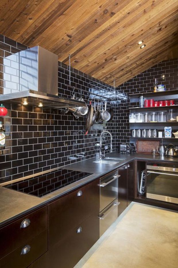 Compact, highly detailed kitchen with dark brown cabinetry, stainless-steel appliances and benchtops, glossy black subway-tile splashback walls, and an exposed timber-plank sloped ceiling. Open shelving displays glassware and cookware, while hanging pots and utensils emphasize the practical industrial character. The space is warmly lit with recessed ceiling lights and under-cabinet lighting.