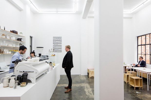 Bright minimalist café interior with a white service counter and commercial espresso machine on the left, open shelving and barista workspace behind it, and a small dining area with timber tables and stools on the right. White walls, exposed structural columns, linear ceiling lighting, a large black-framed window, and a polished industrial concrete floor create a clean contemporary atmosphere.