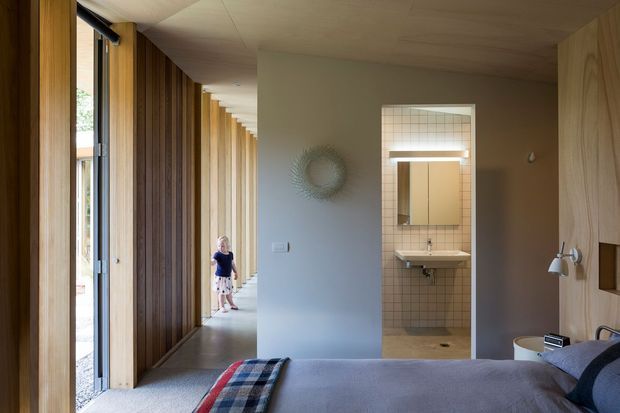 A warm, minimalist bedroom viewed toward an open bathroom doorway. The room features a low-profile bed with muted bedding, extensive vertical timber wall panelling and full-height glazed openings along a corridor, a smooth neutral wall, and a simple wall-mounted reading light. The bathroom beyond has small square tiled walls, a wall-mounted basin, mirror, and shower area. Natural light creates a calm, understated atmosphere.